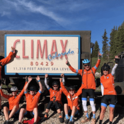 Sarah Zenner and her team share their Moments of Victory at the Climax of Colorado. They are using Davis Phinney's Victory Arms. They are wearing orange bike jerseys, bike pants, and helmets. There is bike on the left side of the picture.