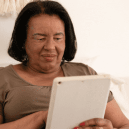 A black woman, middle-aged, looks at an iPad. She has chin-length black hair, red lipstick, and a black shirt. Her iPad case is white. She has red nail polish and is sitting on a white couch with a white wall behind her.