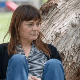 A white woman with shoulder-length brown hair and bangs is sitting in front of a tilted tree. She looks contemplative and sad. She is wearing a black cardigan, a vertical-striped black and white button-down, and blue jeans. There is a playground in the background.