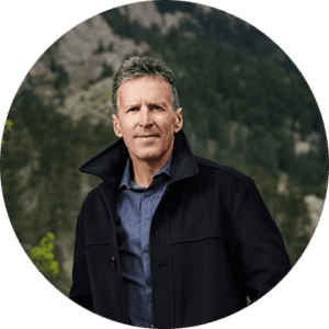 Davis Phinney stands in front of a mountain covered in trees. He is wearing a black coat and a denim button-down. He has short, grey hair and a stoic look on his face.