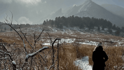 help A woman walking through a field towards snow-covered mountains. It is sunny, and she is bundled up.