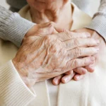Closeup of hands wrapped around an elderly loved one from behind and resting on their heart.